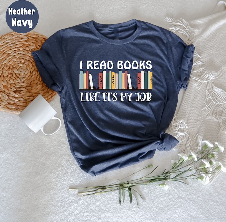 May include: Navy blue t-shirt with the slogan "I READ BOOKS LIKE IT'S MY JOB" in white. The design features a row of colourful books. A white mug and woven basket are in the background.