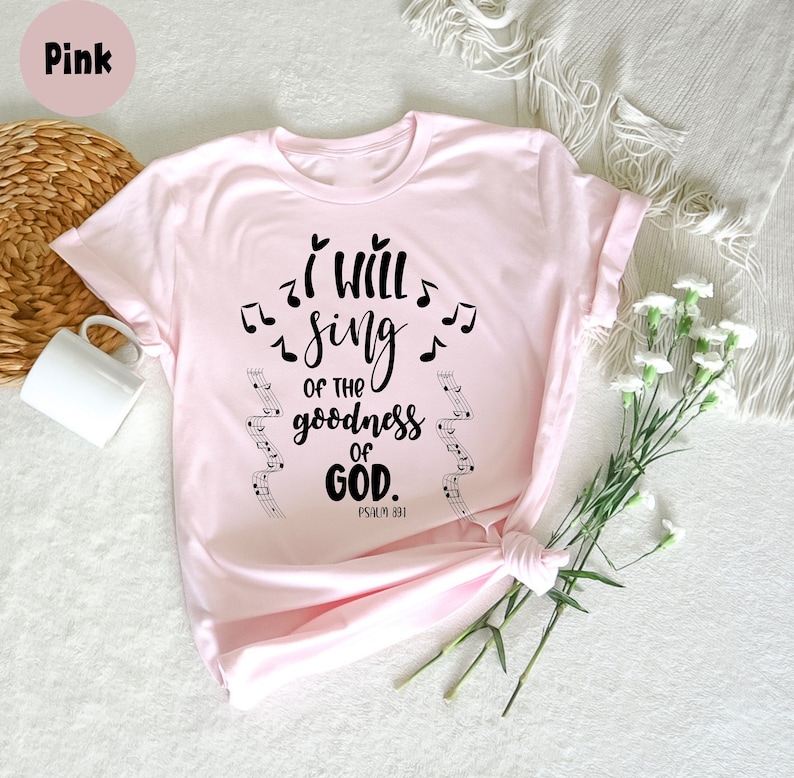 K&ouml;nnte beinhalten: Hellrosa T-Shirt mit dem schwarzen Schriftzug "I will sing of the goodness of God", begleitet von Noten. Das Shirt zeigt auch den Bibelvers Psalm 89,1.