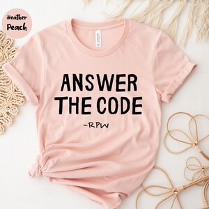 以下が含まれることがあります： 「ANSWER THE CODE」のテキストが黒い太字でプリントされた、ヘザーピーチ色のTシャツ。テキストの下には「-RPW」と書かれています。丸首で袖がロールアップされています。柔らかい素材で作られたシャツです。