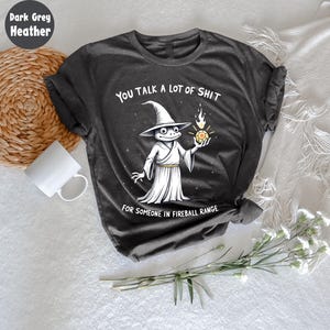 May include: Dark grey heather t-shirt with a humorous graphic. The design features a cartoon frog wizard holding a fireball, with the text "YOU TALK A LOT OF SHIT FOR SOMEONE IN FIREBALL RANGE."