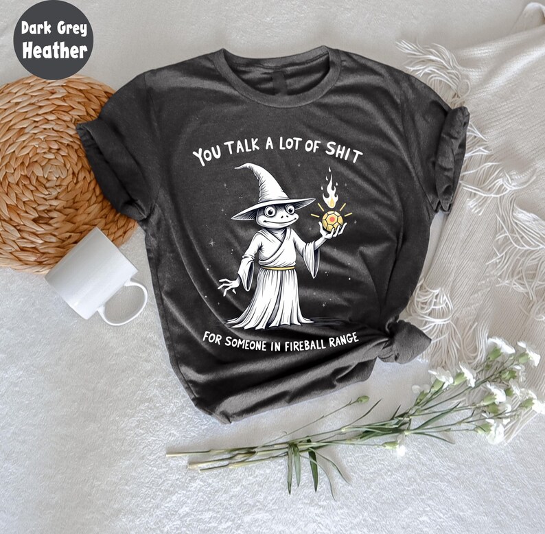 May include: Dark grey heather t-shirt with a humorous graphic. The design features a cartoon frog wizard holding a fireball, with the text "YOU TALK A LOT OF SHIT FOR SOMEONE IN FIREBALL RANGE."