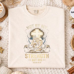 K&ouml;nnte beinhalten: Cremefarbenes Sweatshirt mit einem goldenen und blauen Grafikdesign. Das Design zeigt ein Kreuz, Hanteln und den Text "NOT BY MY STRENGTH BUT HIS". Das Sweatshirt ist von dekorativen Gegenst&auml;nden umgeben.