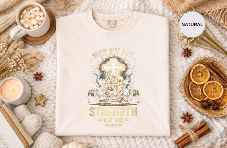 K&ouml;nnte beinhalten: Cremefarbenes Sweatshirt mit einem goldenen und blauen Grafikdesign. Das Design zeigt ein Kreuz, Hanteln und den Text "NOT BY MY STRENGTH BUT HIS". Das Sweatshirt ist von dekorativen Gegenst&auml;nden umgeben.