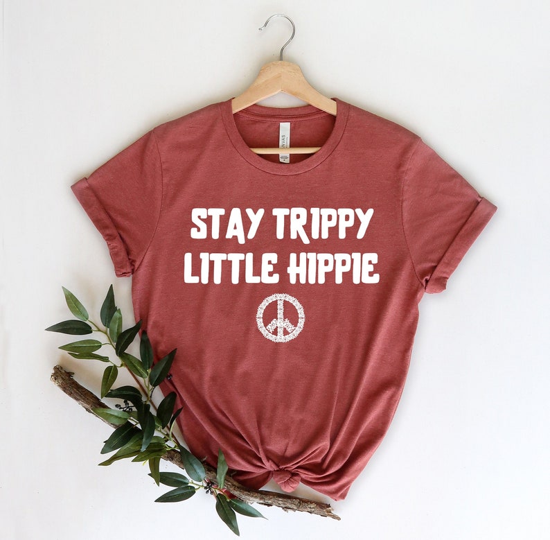 以下が含まれることがあります： 袖をまくったレンガ色のTシャツで、白い文字で「STAY TRIPPY LITTLE HIPPIE」と書かれています。その下に白い平和のシンボルがあります。Tシャツは木製のハンガーに吊るされています。