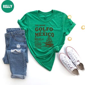 Op de afbeelding: Groen T-shirt met de tekst "The Name Golfo de Mexico" in bruine letters. De tekst "Gulf of Mexico Appeared for the First Time in a World Map in 1550" is ook gedrukt op het T-shirt in bruine letters. Onder de tekst is een schip afgebeeld.