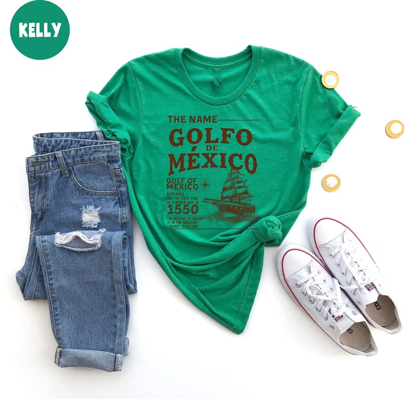 Op de afbeelding: Groen T-shirt met de tekst "The Name Golfo de Mexico" in bruine letters. De tekst "Gulf of Mexico Appeared for the First Time in a World Map in 1550" is ook gedrukt op het T-shirt in bruine letters. Onder de tekst is een schip afgebeeld.
