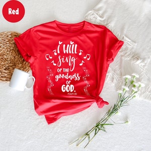 K&ouml;nnte beinhalten: Ein rotes T-Shirt mit dem wei&szlig;en Text "I will sing of the goodness of GOD", Noten und Notenlinien. Das T-Shirt ist am Saum geknotet. Eine wei&szlig;e Tasse und Blumen sind im Hintergrund.