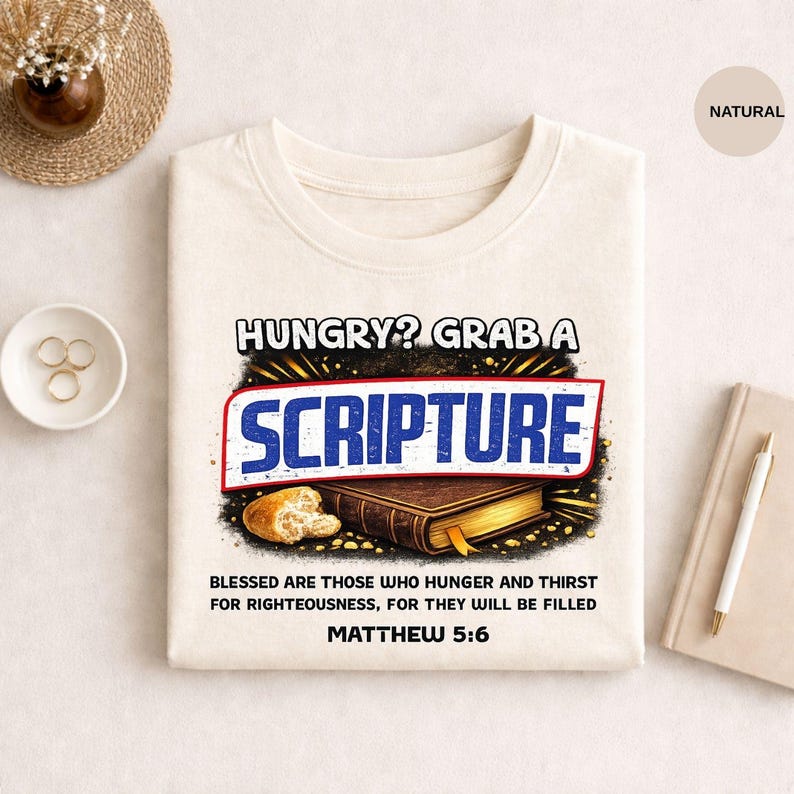 laid flat on a light surface with a centered graphic design. The design features the phrase “HUNGRY? GRAB A” in bold white uppercase letters at the top. Below it, the word “SCRIPTURE” appears in large blue distressed block lettering inside a red outlined shape. Under the text is an illustration of a closed brown Bible with visible pages and a small piece of bread beside it. Beneath the illustration is smaller black text reading “BLESSED ARE THOSE WHO HUNGER AND THIRST FOR RIGHTEOUSNESS