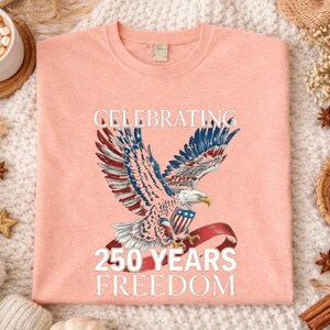 May include: Heather peach-colored t-shirt featuring a detailed illustration of an eagle with outstretched wings, holding a banner with the American flag. The text "CELEBRATING 250 YEARS FREEDOM" is printed above and below the eagle.