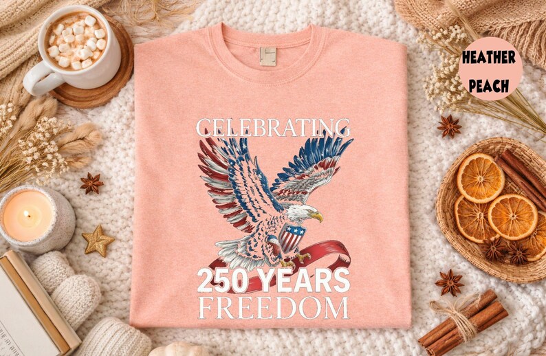 May include: Heather peach-colored t-shirt featuring a detailed illustration of an eagle with outstretched wings, holding a banner with the American flag. The text "CELEBRATING 250 YEARS FREEDOM" is printed above and below the eagle.
