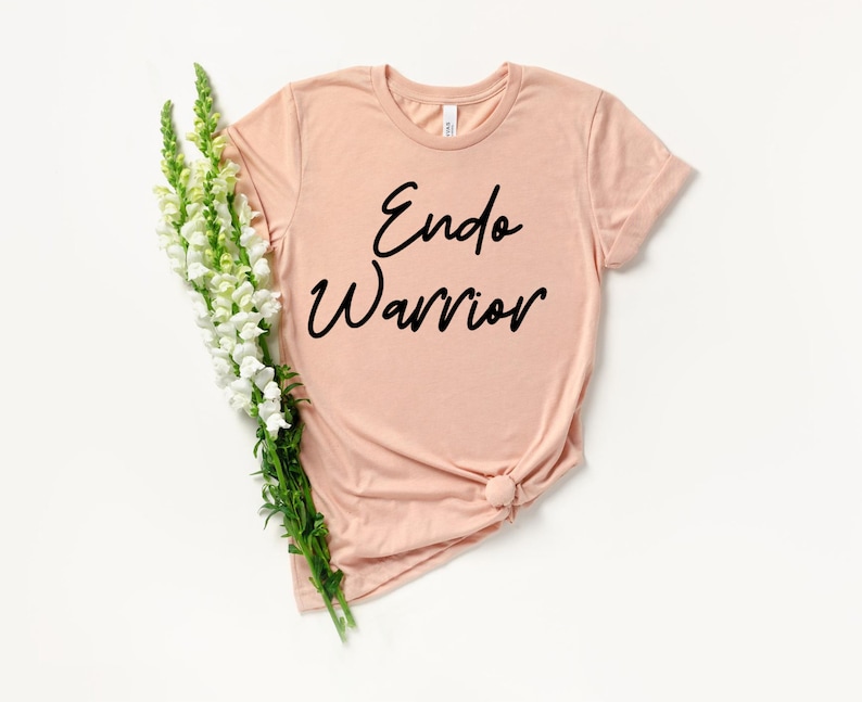 May include: A light peach-colored t-shirt with the words "Endo Warrior" in black script. The shirt is styled with a knot at the bottom and is accompanied by a bouquet of white and green flowers. The t-shirt is made of soft material.