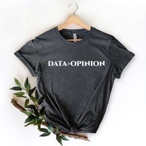 May include: Charcoal grey t-shirt featuring the white text "DATA > OPINION". The garment is displayed on a wooden hanger. A sprig of green foliage is positioned on the lower left against a white backdrop.