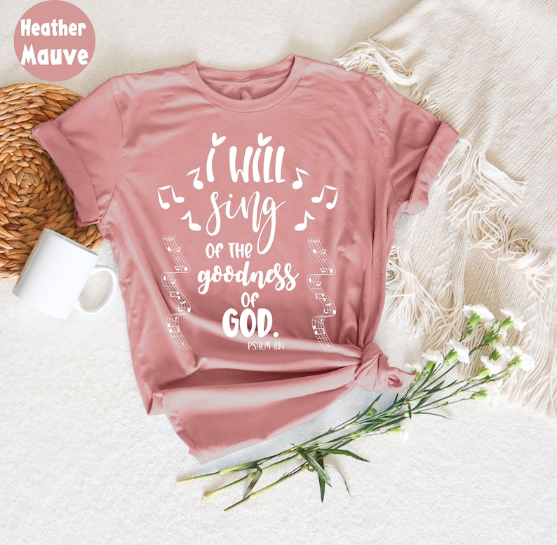 K&ouml;nnte beinhalten: Heather-Mauve-T-Shirt mit dem Text "I WILL SING of the goodness of GOD." Wei&szlig;e Noten und Notenlinien umgeben den Text. Psalm 89:1 ist darunter gedruckt. Eine wei&szlig;e Tasse und Blumen sind im Hintergrund.