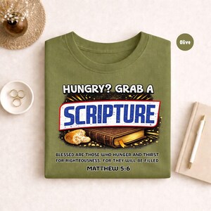May include: Olive green t-shirt with the text "HUNGRY? GRAB A SCRIPTURE" in white and blue. The design includes an image of a book and bread, with the Bible verse Matthew 5:6. A casual, faith-based tee.