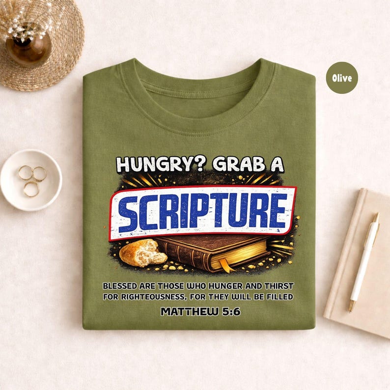 May include: Olive green t-shirt with the text "HUNGRY? GRAB A SCRIPTURE" in white and blue. The design includes an image of a book and bread, with the Bible verse Matthew 5:6. A casual, faith-based tee.