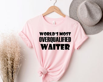 May include: A light pink t-shirt with the text "WORLD'S MOST OVERQUALIFIED WAITER" in bold black letters. The shirt is hanging on a wooden hanger against a white background, with a gray hat visible in the background.