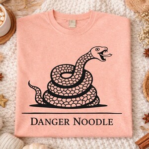 Peut inclure: T-shirt p&ecirc;che avec un motif de serpent noir enroul&eacute; et le texte "DANGER NOODLE". Le serpent a la langue sortie. Le t-shirt est pr&eacute;sent&eacute; sur une surface blanche textur&eacute;e. Le t-shirt est de couleur p&ecirc;che.