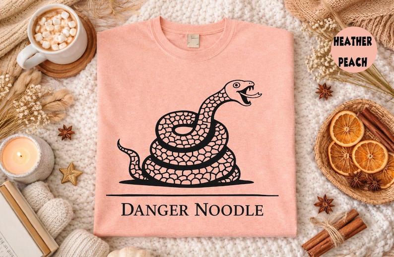 Peut inclure: T-shirt p&ecirc;che avec un motif de serpent noir enroul&eacute; et le texte "DANGER NOODLE". Le serpent a la langue sortie. Le t-shirt est pr&eacute;sent&eacute; sur une surface blanche textur&eacute;e. Le t-shirt est de couleur p&ecirc;che.