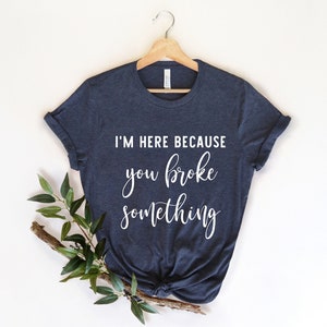 May include: A navy blue t-shirt with the text "I'M HERE BECAUSE you broke something" in white script. The shirt is hanging on a wooden hanger, with a sprig of greenery to the side.