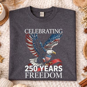 May include: Dark heather gray t-shirt featuring a patriotic design. The graphic includes a bald eagle with red, white, and blue wings, a shield, and the text "CELEBRATING 250 YEARS FREEDOM". The shirt is displayed on a cozy, textured surface.