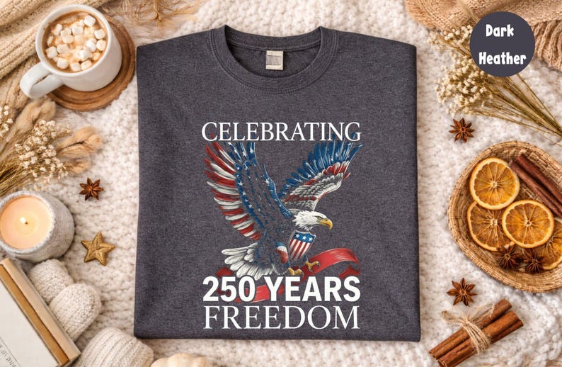 May include: Dark heather gray t-shirt featuring a patriotic design. The graphic includes a bald eagle with red, white, and blue wings, a shield, and the text "CELEBRATING 250 YEARS FREEDOM". The shirt is displayed on a cozy, textured surface.