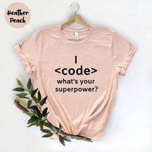 Könnte beinhalten: Hellrosa T-Shirt mit dem Text "I <code/> what's your superpower?" in Schwarz. Das T-Shirt hängt an einem Holzbügel. Die Worte "Heather Peach" befinden sich in der oberen linken Ecke.