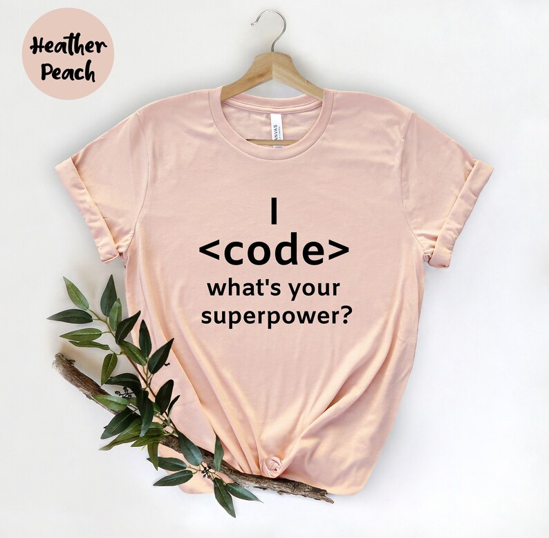 May include: A light pink t-shirt with the text "I <code/> what's your superpower?" in black. The shirt is hanging on a wooden hanger. The words "Heather Peach" are in the top left corner.