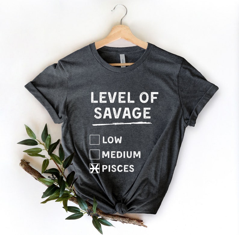Op de afbeelding: Donkergrijs t-shirt met de witte tekst "LEVEL OF SAVAGE". Daaronder staan drie selectievakjes, waarvan de laatste is aangevinkt en is gelabeld met "PISCES". Het t-shirt hangt aan een houten hanger.