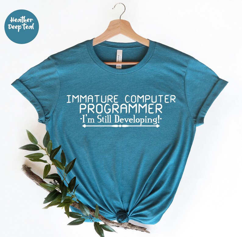 May include: Teal t-shirt with the text "IMMATURE COMPUTER PROGRAMMER I'm Still Developing!" in white. The shirt is hanging on a wooden hanger. The words "Heather Deep Teal" are in a circle in the upper left corner.