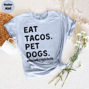 Op de afbeelding: Een heide mint grijs t-shirt met de tekst "EAT TACOS. PET DOGS. #tacos&wigglebutts" in zwart gedrukt.