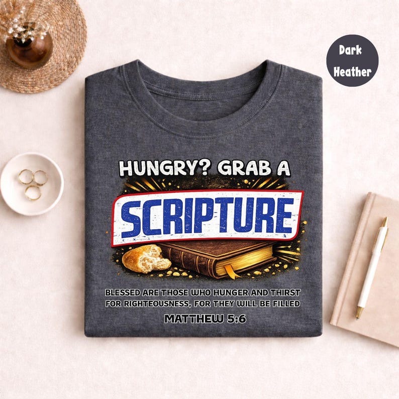 May include: Dark heather t-shirt with the text "HUNGRY? GRAB A SCRIPTURE" in white and blue. The design includes an illustration of a book and bread. The text "BLESSED ARE THOSE WHO HUNGER AND THIRST FOR RIGHTEOUSNESS, FOR THEY WILL BE FILLED MATTHEW 5:6" is also included.