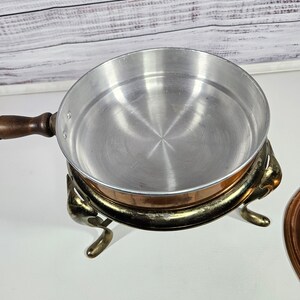 May include: A copper chafing dish with a silver-colored interior and a wooden handle. The dish sits on a decorative stand with three legs.