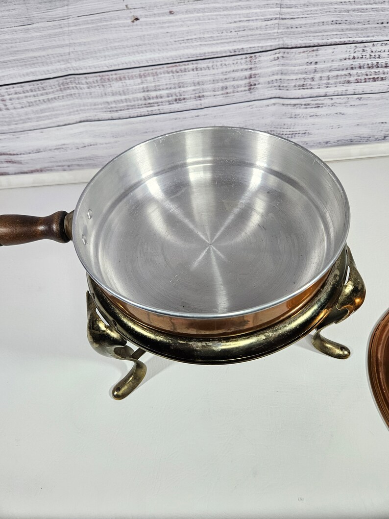 May include: A copper chafing dish with a silver-colored interior and a wooden handle. The dish sits on a decorative stand with three legs.