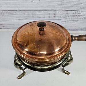 May include: Copper chafing dish with a lid and a stand with three legs. The chafing dish is on a white surface.