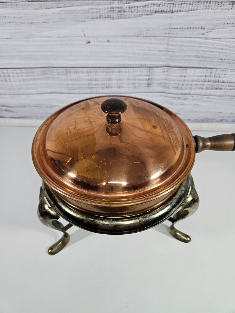 May include: Copper chafing dish with a lid and a stand with three legs. The chafing dish is on a white surface.