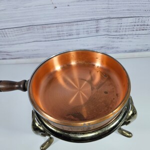 May include: A copper fondue pot with a wooden handle and three decorative legs. The pot is sitting on a white surface.