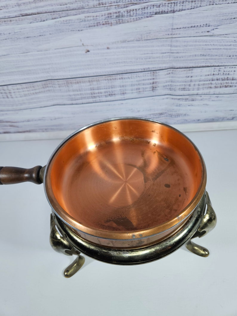 May include: A copper fondue pot with a wooden handle and three decorative legs. The pot is sitting on a white surface.
