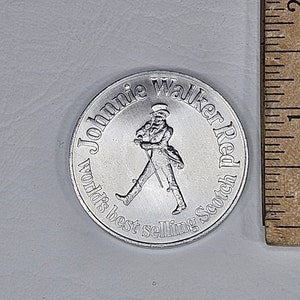 May include: A silver-colored metal token featuring a man in a top hat walking with a cane. The text around the man reads "Johnnie Walker Red World's Best Selling Scotch".