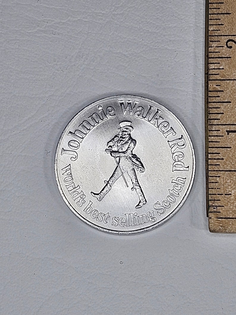May include: A silver-colored metal token featuring a man in a top hat walking with a cane. The text around the man reads "Johnnie Walker Red World's Best Selling Scotch".