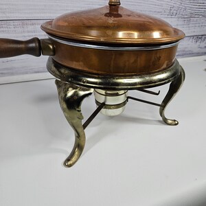 May include: A copper chafing dish with a wooden handle and a gold-tone stand with three legs. The dish has a lid with a knob on top.