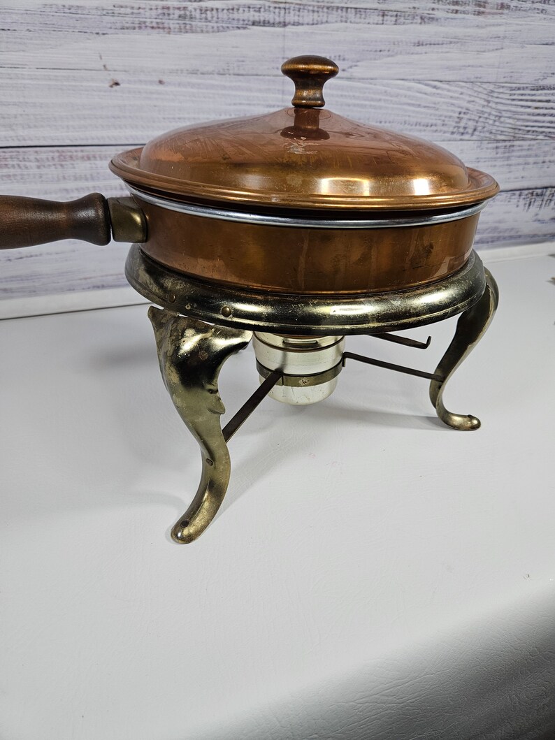 May include: A copper chafing dish with a wooden handle and a gold-tone stand with three legs. The dish has a lid with a knob on top.
