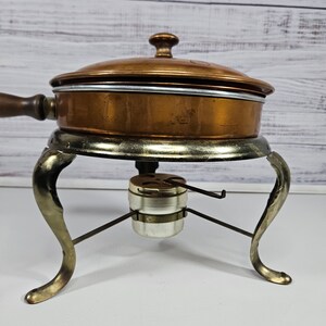 May include: A vintage copper chafing dish with a lid and a wooden handle. The dish sits on a metal stand with three legs and a burner.