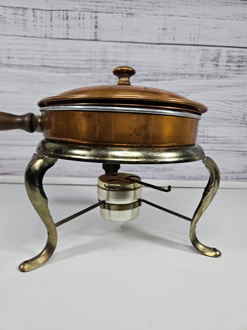 May include: A vintage copper chafing dish with a lid and a wooden handle. The dish sits on a metal stand with three legs and a burner.