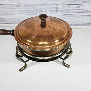 May include: A copper chafing dish with a wooden handle and a lid. The chafing dish is on a silver stand with three legs.