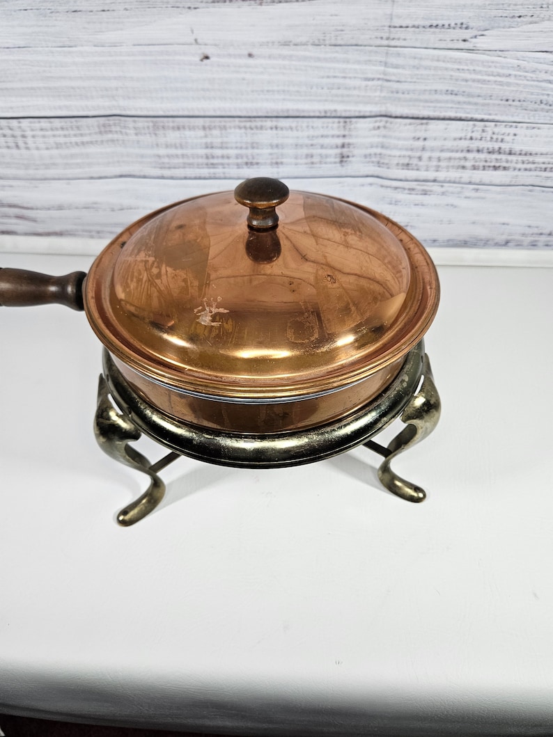 May include: A copper chafing dish with a wooden handle and a lid. The chafing dish is on a silver stand with three legs.