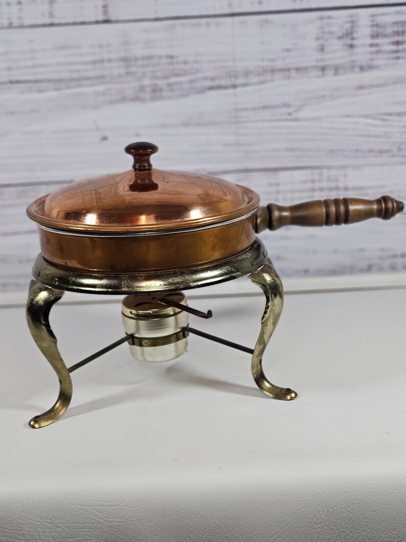 May include: A copper chafing dish with a wooden handle and a silver stand with three legs. The chafing dish has a lid with a knob on top.