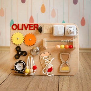 May include: A wooden busy board with various interactive elements for toddlers. Features include gears, a spinner, a bell, an abacus, a shoelace, light switches, and a plug. The name "OLIVER" is displayed in red letters.