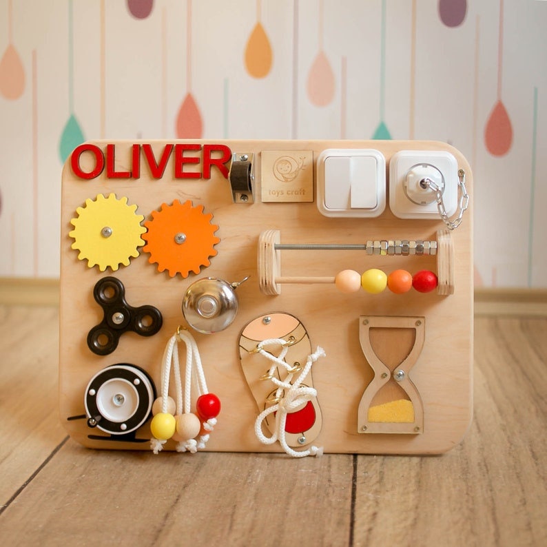 May include: A wooden busy board with various interactive elements for toddlers. Features include gears, a spinner, a bell, an abacus, a shoelace, light switches, and a plug. The name "OLIVER" is displayed in red letters.
