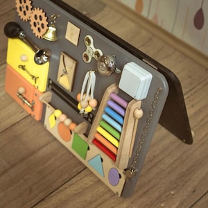 May include: A wooden activity board for toddlers with a variety of textures and activities, including a zipper, a light switch, a spinning gear, a xylophone, a chain, and a bell. The board is painted in various colours, including grey, yellow, orange, and blue.