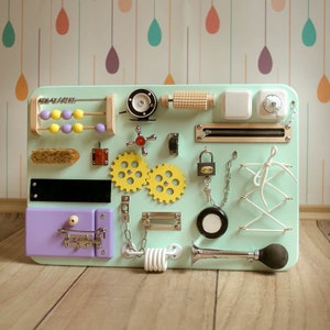 May include: A mint green wooden activity board for toddlers, featuring various interactive elements. Includes an abacus, gears, switches, a zipper, a lock, a horn, and other tactile components. The board is designed to stimulate sensory exploration and fine motor skills.
